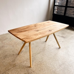 May include: A light wood dining table with a curved edge and three legs. The table is made of natural wood with a light brown finish.