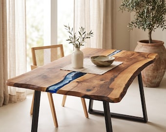 May include: A rectangular dining table featuring a natural live-edge wood slab surface with a central blue resin inlay. The table is supported by black metal U-shaped legs and holds a white ceramic vase with greenery and a shallow ceramic bowl.