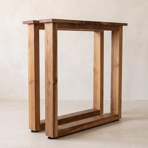 May include: A pair of rectangular table legs constructed from natural brown wood with a visible grain pattern. Each leg features a sturdy frame design with four vertical posts connected by horizontal bars at the top and bottom, finished with small black feet.