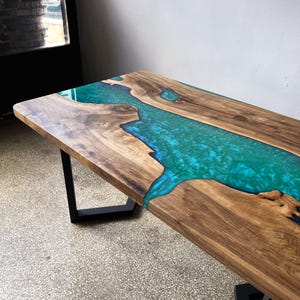 Può includere: Tavolo in legno fatto a mano con un fiume di resina epossidica turchese al centro. Il tavolo ha venature del legno marrone scuro e gambe in metallo nero. La resina epossidica è lucida e riflette la luce. Lunghezza: circa 150 cm.