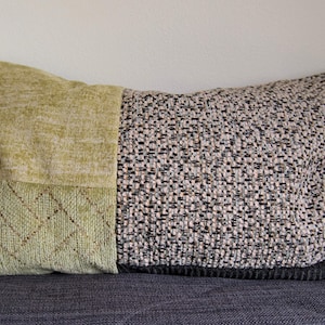 Peut inclure: Un coussin rectangulaire en tissu à motifs verts et noirs. Le coussin est composé de deux tissus différents, l'un à tissage texturé et l'autre à motifs en losange. Le coussin est posé sur une surface gris foncé.