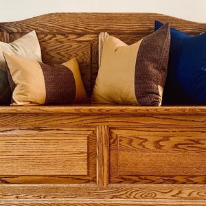 May include: A wooden storage bench with a cushioned top. The bench has a light brown finish and is decorated with four throw pillows. The pillows are in various colours and patterns, including green, brown, beige, and blue.