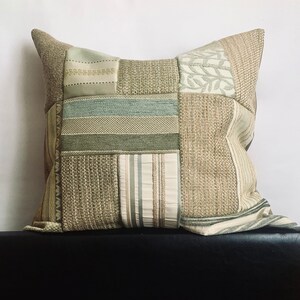 May include: A square cushion with a patchwork design featuring various shades of green and beige fabrics. The cushion has a subtle textured pattern and a simple, understated design.