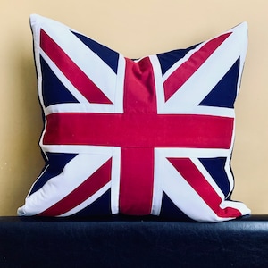 Pode incluir: Uma almofada quadrada com um design Union Jack vermelho, branco e azul. A almofada é feita de tecido e tem uma textura macia e felpuda.