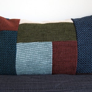 May include: A rectangular cushion with a patchwork design. The cushion is made of different coloured fabrics, including dark blue, light blue, green, brown, and beige. The cushion is sitting on a dark grey surface.