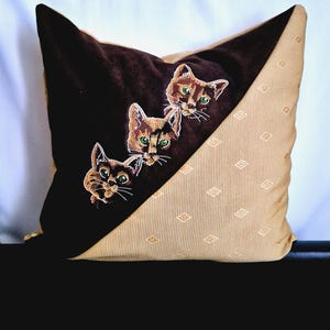 Peut inclure: Un oreiller brun et beige avec trois chats brodés qui regardent par-dessus un rabat brun. Les chats ont des yeux verts et une fourrure brune.