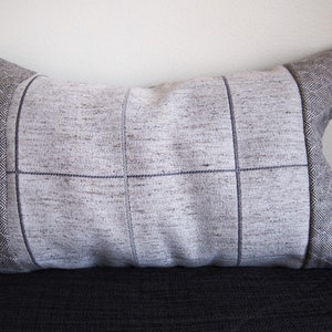 May include: Rectangular cushion with a light grey woven fabric centre, featuring a grid pattern created by dark blue stitching. The cushion ends have a grey herringbone pattern. The cushion is resting on a dark surface.