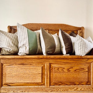 May include: A wooden storage bench with a cushioned top and four throw pillows. The pillows are striped in beige and white, with two additional pillows in green and brown.