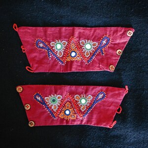 Peut inclure: Deux manchettes en tissu rouge avec broderies complexes. Les manchettes présentent des broderies colorées en bleu, orange et blanc, avec de petits miroirs et des boutons en bois. Elles font probablement partie d'un vêtement traditionnel.