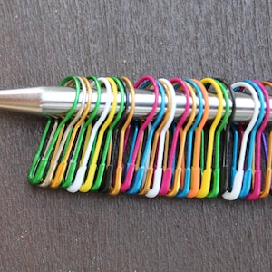 Op de afbeelding: Een verzameling kleurrijke steekmarkeerders in verschillende tinten, waaronder groen, geel, roze en blauw, is bevestigd aan een zilveren breinaald. De markeerders zijn gemaakt van metaal en zijn in een rij gerangschikt, waardoor een levendige weergave ontstaat.