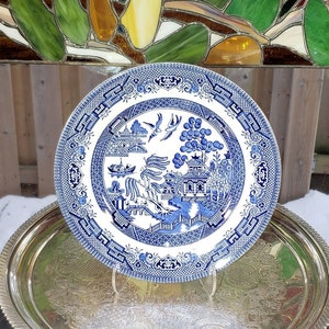 May include: A blue and white porcelain dinner plate featuring a classic willow pattern design. The plate depicts a stylised landscape with a pagoda, trees, and a bridge.