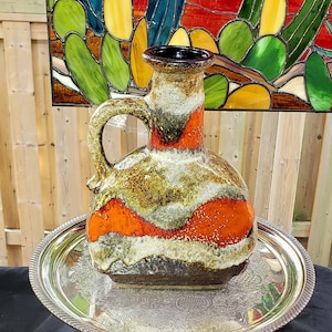 May include: A ceramic vase with a brown, white, and orange glaze. The vase has a wide base and a narrow neck. It is sitting on a silver tray.