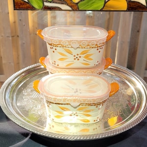 May include: Three ceramic casserole dishes with lids. The dishes are white with a brown and orange floral design. The lids are clear plastic. The dishes are stacked on a silver tray.