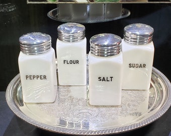 May include: Four white ceramic shakers with silver lids on a silver tray. The shakers are labeled "Pepper", "Flour", "Salt", and "Sugar".