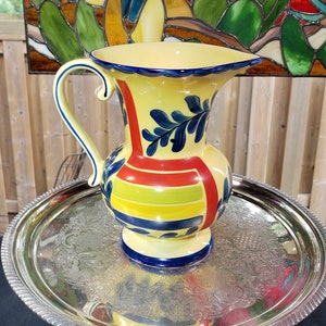 May include: A yellow ceramic pitcher with blue and red painted designs sits on a silver tray. The pitcher has a wide mouth and a handle.