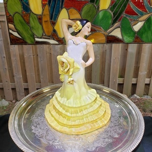 May include: A porcelain figurine of a woman in a yellow dress with a large yellow rose on her hip. The figurine is standing on a silver tray.