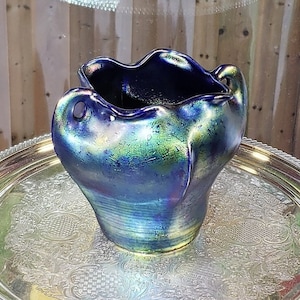 May include: A small, iridescent blue vase with a ruffled rim. The vase has a unique shape and a glossy finish, with hints of green and gold. It sits on a silver tray with an ornate pattern.