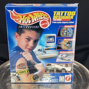 Op de afbeelding: Een blauwe Hot Wheels Tattoo Designer CD-ROM-doos met een jongen met een tijdelijke tatoeage. De doos bevat een computergrafiek, een witte Hot Wheels-auto en de tekst "Maak aangepaste tijdelijke tatoeages!"
