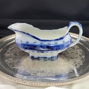 May include: A white ceramic gravy boat featuring a blue transferware pattern with floral and geometric motifs. The vessel has a curved handle, a scalloped rim, and a pouring spout, resting on a silver-toned metal tray with an ornate embossed design.