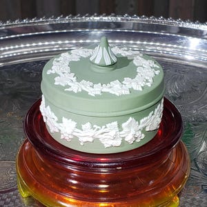 May include: A small, round, sage green trinket box with a white floral relief design. The lid has a conical finial. The box sits on a layered, amber-coloured glass stand, against a silver tray background.