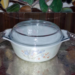 May include: A vintage Pyrex casserole dish with a clear glass lid. The off-white dish features a floral design in blue and orange. The dish has two handles and is sitting on a silver tray.