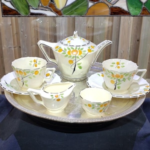 May include: A cream-coloured ceramic tea set featuring a teapot, two teacups with matching saucers, a creamer, and a sugar bowl. Each piece is decorated with a hand-painted pattern of orange fruit and green leaves with black accents on a silver tray.