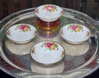 May include: A large, round, polished silver-tone serving tray with an embossed border, holding four small, white porcelain bowls. Each bowl features a delicate floral pattern of pink and yellow roses with green leaves and a gold-tone rim.