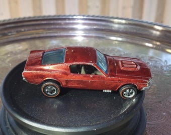 May include: A metallic red die-cast toy car featuring a fastback coupe body style, a bonnet scoop, and four wheels with redline tyres. The vehicle has a visible interior, clear windows, and silver-coloured exhaust pipes along the side panels.