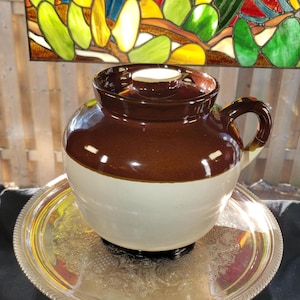 May include: A two-toned ceramic crock with a dark brown glazed upper section and a cream-coloured lower body. The vessel features a matching brown lid with a central knob and a single side handle, resting on a round, ornate silver-toned metal tray.
