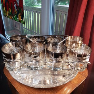 May include: A set of eight clear glass tumblers with silver metallic rims and etched floral patterns, arranged in a silver-toned metal wire carrier that sits atop a matching round silver-toned metal serving tray with an embossed border.