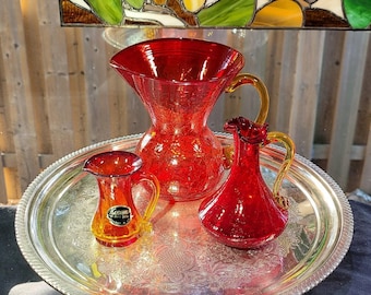 May include: A set of three red crackle glass vessels with amber-colored handles, consisting of a large pitcher and two smaller pitchers, arranged on a round, ornate silver-toned metal tray with an embossed floral pattern and a rope-style edge.