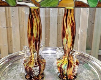 May include: A pair of tall glass vases featuring a swirling marbled pattern of brown, yellow, and cream colors. Each vase has a slender neck, a rounded base, and two clear glass decorative handles attached to the sides with textured glass details.