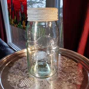 May include: A clear glass mason jar featuring an embossed beaver graphic on the front and the word BEAVER embossed below it. The jar is topped with a textured, light-coloured metal screw-on lid with a weathered, matte finish and ridges.