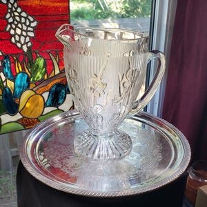 May include: A clear glass pitcher featuring a vertical ribbed texture and embossed floral iris motifs, resting on a round silver-toned metal serving tray with an intricate engraved pattern along the center and a decorative rope-style edge.
