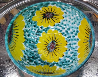 May include: A round ceramic bowl featuring a vibrant pattern of large yellow flowers with dark centers. The background is filled with a textured teal and light blue dotted design. The bowl has a smooth, glossy finish and a wide, circular rim.