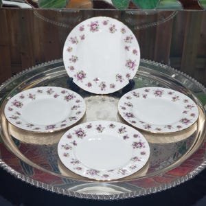 May include: A set of four white ceramic plates featuring a delicate floral pattern with small pink and purple flowers and green leaves around the rim. Each plate has a smooth, circular design with a thin gold-toned edge, arranged on a silver tray.