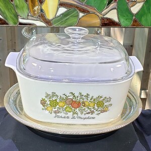May include: A white rectangular ceramic casserole dish with a clear glass lid and side handles. The front features a colourful botanical illustration of vegetables and herbs with the text L'Echalote La Marjolaine printed in black script below.