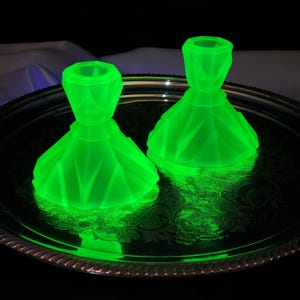 May include: Two vibrant neon green glass candle holders with a faceted, geometric design. Each piece features a wide, angled base that tapers upwards to a cylindrical opening. The glass glows intensely, reflecting light across the surface.