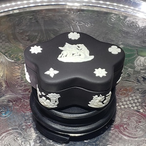 May include: A black, star-shaped trinket box with white relief detailing. The box is decorated with floral and mythological scenes, and the lid shows two animals. It rests on a black, tiered base, presented on a silver tray.