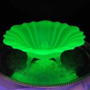May include: A vibrant green glass bowl with a scalloped rim and three small feet. The bowl has a textured, ribbed design and sits on a silver-colored tray. The bowl's color is a bright, almost neon, green.