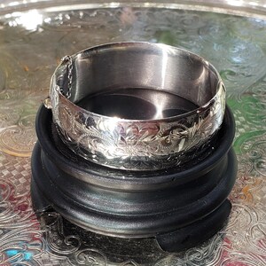 May include: A silver bangle bracelet with an engraved floral design. The hinged bracelet has a safety chain. It is displayed on a black, round stand, against a reflective, patterned surface.
