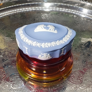 May include: A pale blue ceramic trinket box with white relief detailing, featuring figures and a leaf border. The box is placed on a layered, amber-coloured glass container, displayed on a silver tray.