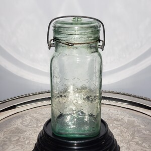 May include: A vintage light green glass mason jar with a wire bail closure and a matching glass lid. The front of the jar features an embossed shield design with the text "WIDE MOUTH" and "ADJUSTABLE" visible on the lower portion of the glass body.
