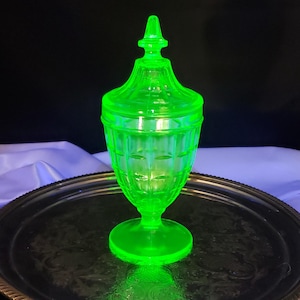 May include: A vibrant green glass jar with a lid, set on a silver tray. The jar has a decorative, geometric pattern and a pedestal base. The lid is topped with a pointed finial. The glass glows brightly, suggesting a vintage or antique piece.