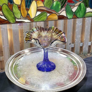 Peut inclure: Une coupe en verre bleu cobalt avec un bord irisé et ondulé ainsi qu'une base texturée et à motifs. La coupe est posée au centre d'un grand plateau de service rond en métal argenté orné d'un motif floral et de volutes en relief.