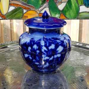 May include: A round, bulbous glass jar featuring a deep cobalt blue and white mottled, cloud-like pattern. The jar includes a matching lid with a small, pointed finial at the top. The glass has a smooth, glossy finish with a scalloped edge.