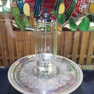 May include: A tall, clear glass canister with a vertical paneled design and a polished chrome lid, featuring a central metal rod and a circular base insert, resting on a large, round, silver-toned metal serving tray with an ornate embossed pattern.