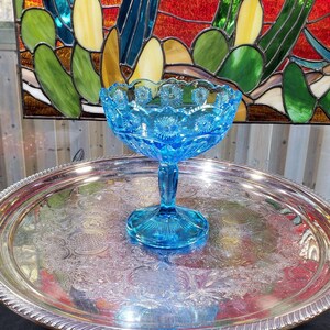 Peut inclure: Une coupe à pied en verre bleu vif avec un bord festonné et un motif de marguerites pressées sur le bol. La base du piédestal est circulaire avec un motif rayonnant en étoile. La coupe est centrée sur un grand plateau rond en métal argenté.