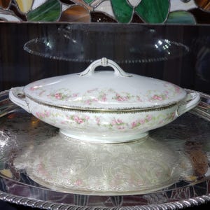 Può includere: Un piatto da portata in porcellana bianca con coperchio, ornato da motivi floreali di rose rosa e finiture dorate. Il piatto ha due manici e un manico sul coperchio. Poggia su un vassoio d'argento dal design ornato.