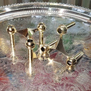 May include: Two polished brass candle holders, each featuring a three-armed design with cylindrical sockets and spherical joints. The metallic finish reflects light, highlighting the smooth, geometric structure of these candle stands.